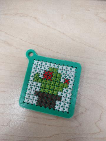 Green 3D printed keychain with cross stitched cactus design. Cactus is green with red flowers, in a brown pot, with a white background.